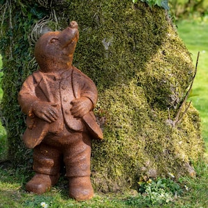 May include: A rustic, metal garden ornament depicting a mole in a suit, with a weathered, brown finish. The mole statue stands in front of a moss-covered tree. A charming, detailed piece for the garden.