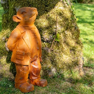 May include: A weathered, rust-colored garden statue of a creature in a suit, standing on grass. The statue has large ears and a smiling expression, with details like a jacket, pockets, and large feet. The statue is positioned next to a moss-covered tree.