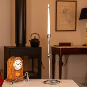 Op de afbeelding: Een zilveren metalen kandelaar met een brandende witte kaars. De kandelaar heeft een knoopontwerp en een cirkelvormige basis. Een houten klok en een open haard zijn op de achtergrond te zien.
