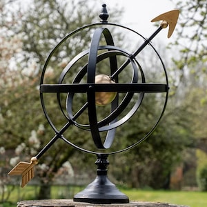 May include: A black metal armillary sphere with a gold-coloured arrow and a central sphere. The decorative garden ornament features intersecting rings and sits on a black pedestal. The arrow points through the sphere.