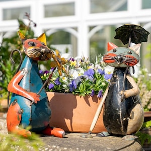 May include: Two metal cat sculptures, one blue and orange, the other black and silver, stand near a flower pot. The blue cat holds a flower, while the black cat has a small umbrella. The cats are whimsical garden decor.