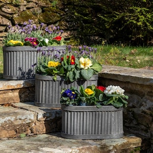 May include: Three grey, ribbed metal planters filled with colourful flowers, including yellow, red, purple, and white blooms. The planters are arranged on stone steps, creating a tiered display. The scene is outdoors.