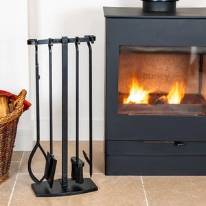 May include: A black fireplace tool set with a stand, including a poker, shovel, brush, and tongs. The tools are positioned in front of a black fireplace with a fire burning inside. A wicker basket with firewood is visible on the left.