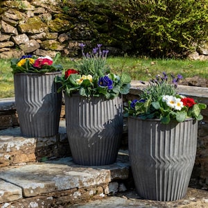 Op de afbeelding: Drie grijze metalen plantenbakken gevuld met kleurrijke bloemen, waaronder rode, gele, blauwe en witte bloemen. De plantenbakken hebben een verticaal gegolfd ontwerp en zijn geplaatst op stenen trappen in een tuin.
