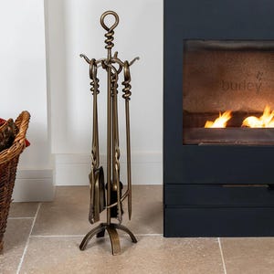 May include: A bronze fireplace tool set with a stand. The set includes a poker, shovel, brush, and tongs. The tools have decorative twisted handles and are arranged on a tripod base. The stand has a looped top. The tools are made of metal.