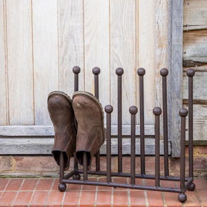 May include: A brown metal boot rack with a series of vertical rods topped with small spheres. Two brown leather boots are placed in the rack. The rack is on a brick surface, with a wooden door in the background.