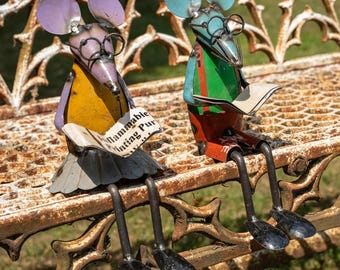 May include: Two whimsical metal mouse sculptures sit on a rusty bench, each wearing glasses and reading. One mouse has purple ears and a yellow top, the other has blue ears and a green top. The text on the paper reads "Flammable Printing Pur".