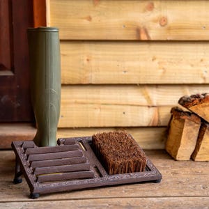 May include: A brown boot scraper with a built-in brush and a green rubber boot. The boot scraper is made of metal and has a decorative design. The brush is made of brown bristles. The background is a wooden wall.