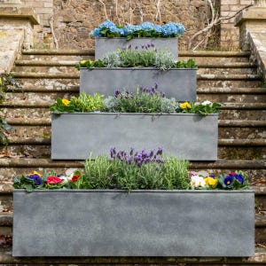 May include: Four grey rectangular planters filled with diverse flowers and greenery, arranged on stone steps. The top planter has blue hydrangeas, while others display lavender, yellow, and red blooms, creating a tiered garden display.