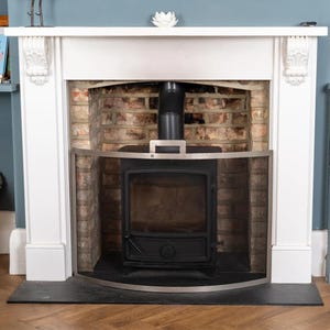 Può includere: Una cornice per camino in legno bianco con mensole decorative sovrasta una stufa a legna in metallo nero. Un parascintille in metallo argentato curvo è posizionato davanti alla stufa, inserita in un focolare rustico in mattoni a vista.