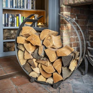Può includere: Un portaleña circolare in metallo pieno di legna tagliata. Il supporto ha un design curvo con una maniglia e poggia su tre piccoli piedi. La legna è un mix di pezzi marroni chiari e scuri.
