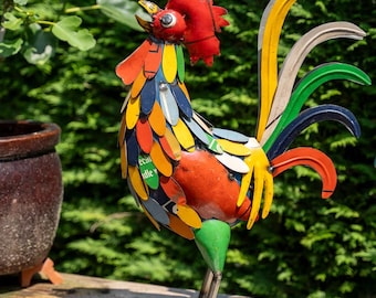 Puede incluir: Escultura de gallo de metal colorido con cresta y barbilla rojas. El cuerpo está hecho de piezas de metal de varios colores. Las plumas de la cola son amarillas, verdes y azules. La escultura está sobre una superficie de madera.