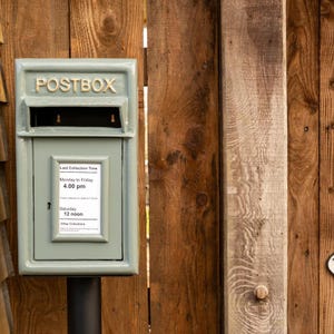May include: A sage green postbox with the word "POSTBOX" in gold lettering. The postbox has a slot for mail and a small door with a collection time schedule. The postbox is mounted on a black post and set against a wooden fence.