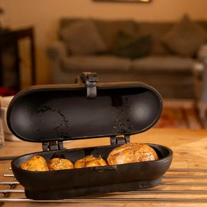 May include: Black cast iron bread baker with a hinged lid, containing four baked rolls. The baker sits on a metal rack over a wooden surface. The background shows a living room with a sofa.