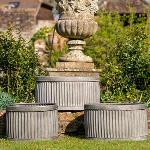 Peut inclure: Trois jardinières en métal gris vieilli avec des détails à nervures verticales. Les jardinières sont de tailles différentes et sont posées sur une pelouse herbeuse devant un mur en pierre et une grande urne en pierre. La scène se déroule à l'extérieur.