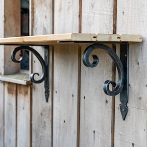 May include: A wooden shelf with two black, wrought iron brackets. The brackets have a decorative scroll design and are mounted on a light-colored wooden wall. The shelf is made of light-colored wood.