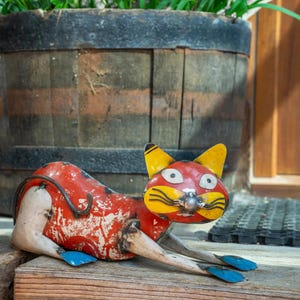 May include: A metal cat sculpture with a red body, yellow ears, and blue feet. The cat is lying down on a wooden surface. The sculpture has a distressed, vintage appearance.