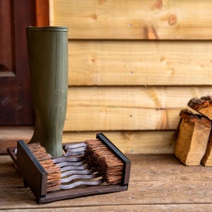 May include: An olive green wellington boot in a brown metal boot cleaner with two brushes. The boot cleaner has a wavy design and is on a wooden surface. Firewood is in the background.