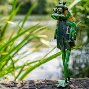May include: A whimsical metal frog sculpture with a green and yellow color scheme. One frog is riding on the back of another, with the sculpture standing on long green legs. The sculpture is set against a blurred background of greenery and water.