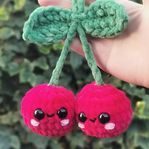 May include: Two crocheted cherries with green stems and leaves. The cherries have smiling faces with black eyes and white accents.