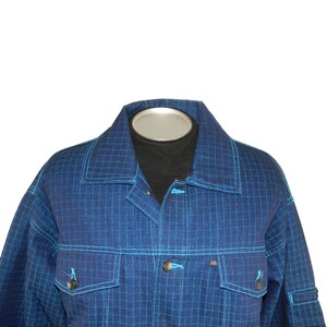 May include: A blue grid-patterned jacket with light blue stitching and accents. The jacket features two front pockets with button closures and a button-down front. The collar and cuffs are also detailed with light blue stitching. The jacket is displayed on a mannequin.