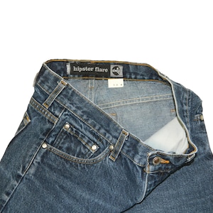 May include: Close-up of blue denim jeans with the text "hipster flare" on the waistband. The jeans have a classic five-pocket design with rivets and a button closure. The denim is a medium wash, and the jeans appear to be unworn.