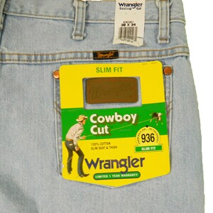 Op de afbeelding: Lichtblauwe denim jeans met een geel en groen Wrangler Cowboy Cut-label. Het label toont een cowboy die een paard berijdt en de tekst "Slim Fit" en "936".