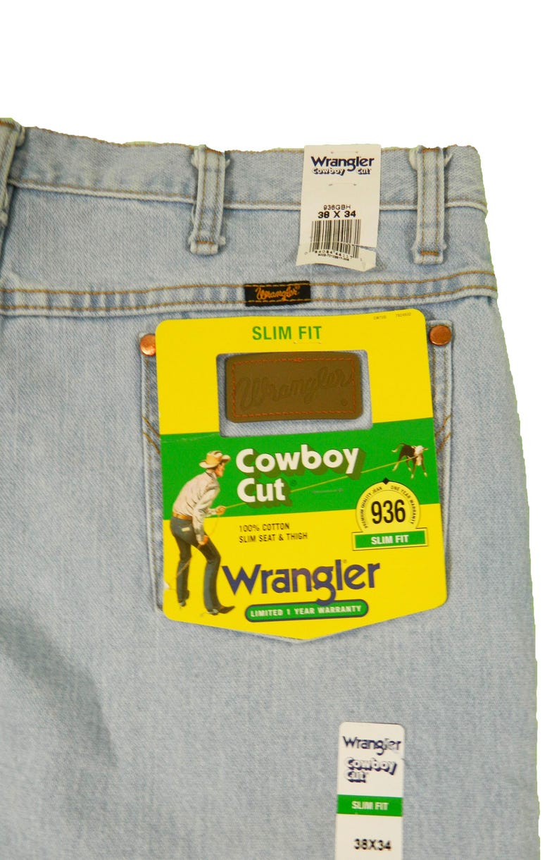 Op de afbeelding: Lichtblauwe denim jeans met een geel en groen Wrangler Cowboy Cut-label. Het label toont een cowboy die een paard berijdt en de tekst "Slim Fit" en "936".