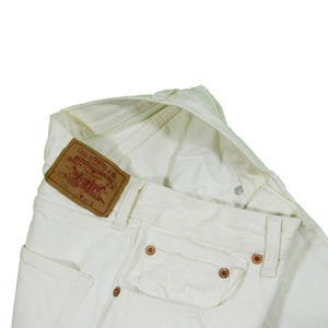 May include: Close-up of off-white denim jeans with a brown leather Levi Strauss & Co. patch. The jeans have copper rivets and a classic five-pocket design. The fabric appears slightly textured.