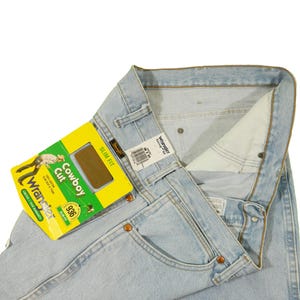 Op de afbeelding: Lichtblauwe denim jeans met een geel en groen label met de tekst "Wrangler Cowboy Cut Slim Fit 936".
