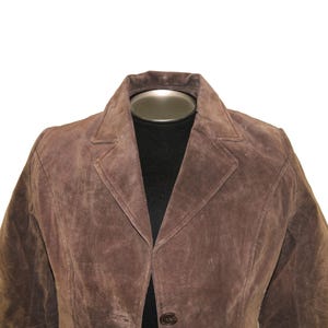 May include: A brown suede blazer with a notched lapel collar and two-button closure. The jacket is displayed on a black mannequin, showcasing its tailored fit and classic design. The blazer's rich color and texture are prominent.