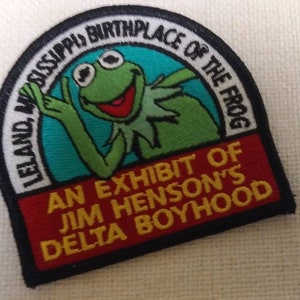 May include: A black and red embroidered patch with a green frog and the text "LELAND, MISSISSIPPI, BIRTHPLACE OF THE FROG AN EXHIBIT OF JIM HENSON'S DELTA BOYHOOD".