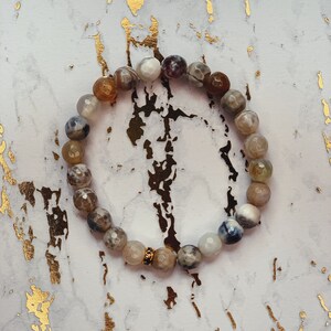 May include: A bracelet made of agate beads in shades of brown, white, and gray. The beads are strung on a clear elastic cord and have a gold-toned accent bead.
