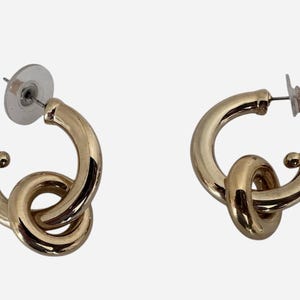 May include: A pair of gold-colored hoop earrings. Each earring features a knot design, with a curved, polished surface. The earrings have post-style backs with clear plastic and metal components.