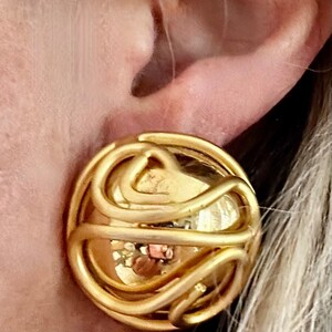 May include: Gold-toned clip-on earring with a circular design. The earring features a rounded base with a swirling, openwork pattern overlaying the surface. The design is a bright, shiny gold color.