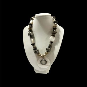 May include: A necklace featuring a series of dark brown wooden beads, metallic silver-toned textured spheres, and smooth white circular shell discs. The centrepiece is a large white shell disc with a smaller, textured silver-toned metal ring.