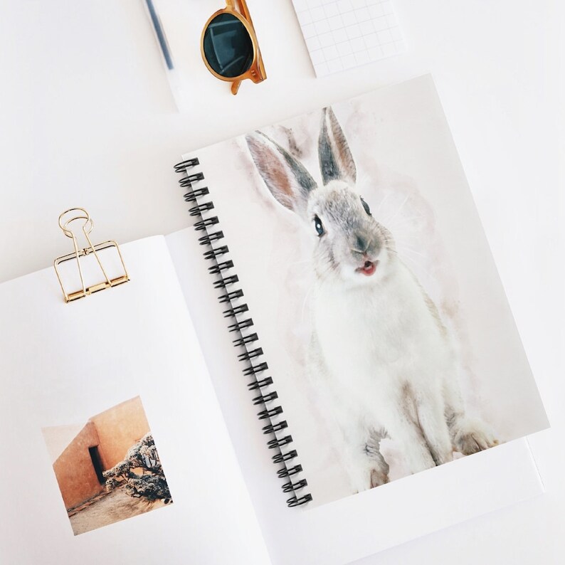 May include: A white rabbit with gray ears and a pink nose is looking directly at the camera with its mouth open. The rabbit is on a white background with a watercolor effect.