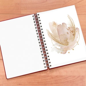 May include: Open spiral-bound notebook with a red cover. The right page features a watercolor floral design in blush pink and gold. The left page is blank. The notebook is on a light brown wooden surface.