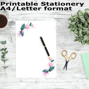 May include: Printable stationery in A4/Letter format with a floral design featuring pink flowers and green leaves. The stationery is on a white background and is surrounded by a wooden surface with a cup of coffee, a pair of scissors, a potted plant, and a green notebook.