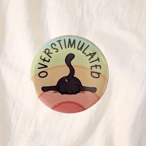 May include: A round button with the word "OVERSTIMULATED" in black text around the edge. A black cat illustration is in the center, with its body partially submerged. The background has a gradient of colors, including green, tan, and pink.