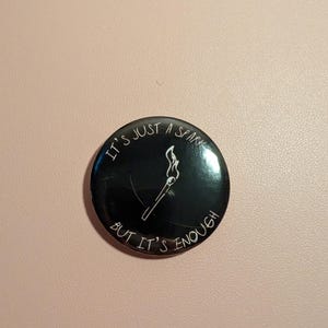 Puede incluir: Un botón negro con texto blanco y una ilustración de cerilla. El texto dice "IT'S JUST A SPARK BUT IT'S ENOUGH" en un círculo. La cerilla está encendida, con una llama en la parte superior.