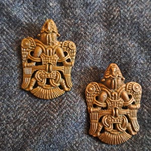 May include: Two identical bronze-coloured metal brooches featuring an intricate, symmetrical relief design of a stylised bird or creature with wings spread, detailed geometric patterns, and a textured surface finish on each piece.