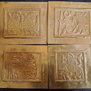 May include: Four square, gold-colored metal plaques with embossed relief designs. Each plaque features a different scene of figures in armor, some on horseback, with spears and shields. The plaques are arranged in a square formation.