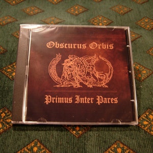 Puede incluir: Una caja de CD con un fondo marrón oscuro y una imagen dorada de una criatura alada. El texto "Obscurus Orbis" y "Primus Inter Pares" está impreso en dorado en la caja.
