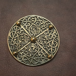 May include: A circular, brass-colored metal ornament with intricate Celtic knotwork designs. The ornament has five raised, round, brass-colored metal studs.
