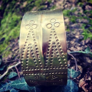 Puede incluir: Un brazalete de metal dorado con un diseño geométrico. El brazalete tiene una superficie texturizada y está decorado con círculos y triángulos.