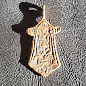 May include: A bronze pendant with an elaborate, symmetrical design. The pendant has a loop at the top for a chain and features a series of vertical and curved lines, creating a detailed pattern. The pendant is approximately 5 cm long.