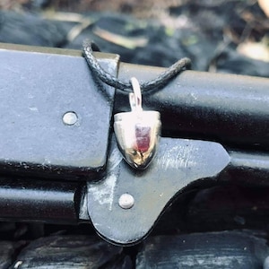May include: A silver bullet charm pendant on a black cord necklace. The pendant is shaped like a bullet and has a shiny, metallic finish.