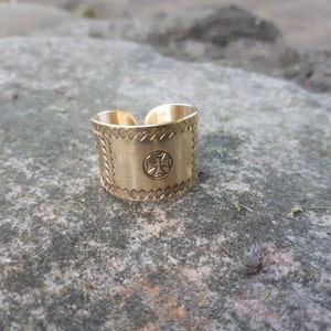 Könnte beinhalten: Ein goldfarbener Metallring mit einem Kreuzdesign und einem strukturierten Muster an den Rändern.