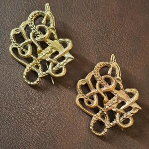 May include: Two gold-toned metal brooches shaped like intertwined snakes. The brooches are detailed and intricate, with each snake's body forming a knot.